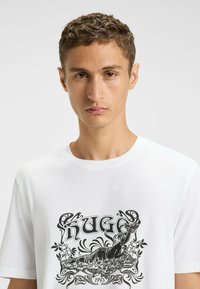 Witte katoenen t-shirt met een zwart grafisch ontwerp dat een vrouw en bloemmotieven toont, met de tekst "HUGO 1993." Klassiek design met rechte hals.