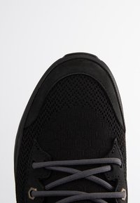 Baskets noires avec tige en mesh et lacets gris foncé noués en motif croisé près de la zone des orteils.