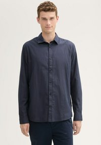 Camisa de botones de manga larga azul marino, hecha de un tejido ligero con una textura suave, que presenta un collar puntiagudo y bolsillos frontales.