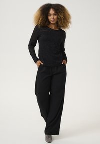 Top in pizzo nero a maniche lunghe con un motivo floreale, abbinato a pantaloni neri a gamba larga e scarpe con tacco alto argento lucido.