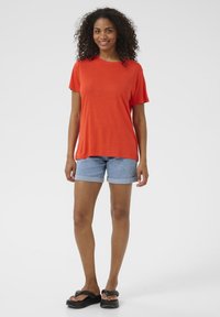 Červené tričko s krátkým rukávem v kombinaci se světle modrými denimovými šortkami se zahnoutavými lemy a černými žabkami. Hladká textura, volný střih.