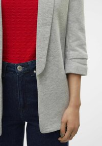Blazer gris clair avec des manches retroussées, associé à un haut texturé rouge et un jean en denim foncé. Détail des boutons en argent notable au niveau de la taille.