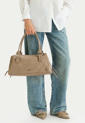 Persona con jeans azul claro y camisa blanca sosteniendo un bolso de cuero taupe con doble asa y correas con hebilla.