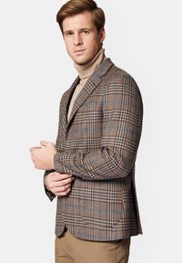 Brązowa marynarka w kratę z niebieskimi akcentami, dopasowany krój, wysoki kołnierz, teksturowany materiał, noszona na beżowym swetrze z golfem.