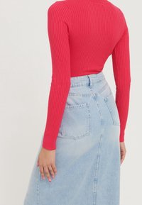 Femme portant un haut rouge à manches longues côtelé, rentré dans une jupe en jean bleu clair taille haute avec des poches arrière, vue de dos.