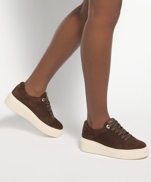 Personne portant des baskets compensées en daim marron avec des semelles blanches, montrant les jambes de genoux vers le bas sur un fond clair uni.