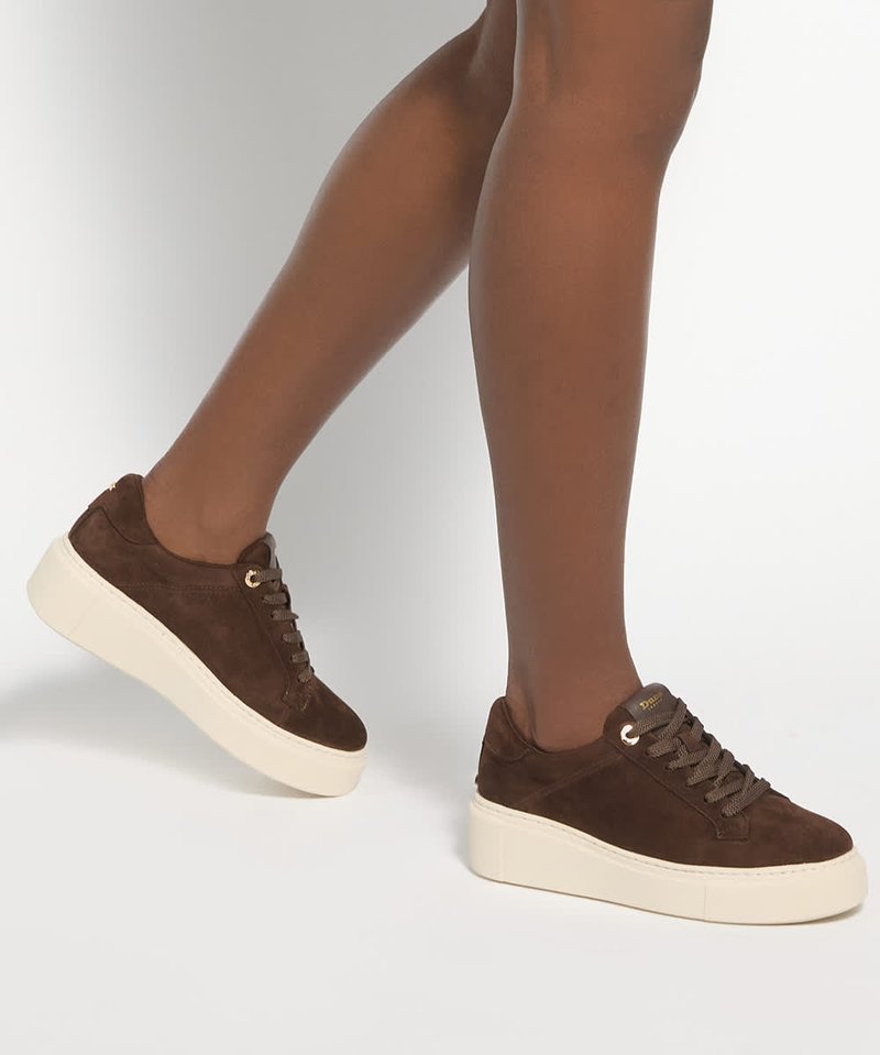 Personne portant des baskets compensées en daim marron avec des semelles blanches, montrant les jambes de genoux vers le bas sur un fond clair uni.