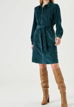 Kobieta ubrana w turkusową sztruksową sukienkę z guzikami i paskiem, zestawioną z jasnobrązowymi skórzanymi kozakami za kolano, stojąca na białym tle.