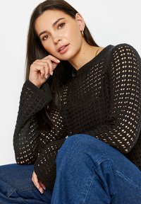 Maglione nero a lavorazione a rete aperta, con colletto e polsini a costine. Abbinato a jeans blu. Le maniche sono leggermente oversize.