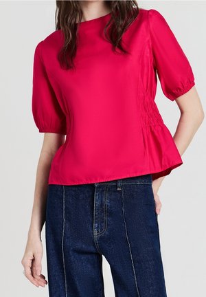 Femme portant un chemisier rouge vif à manches courtes bouffantes et un jean taille haute bleu foncé avec des détails de coutures visibles.