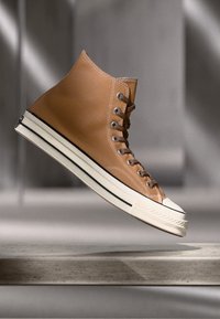 Høye joggesko laget av lys brun lær med hvit gummisåle, svart stripe detalj og snøring med metalløyler.