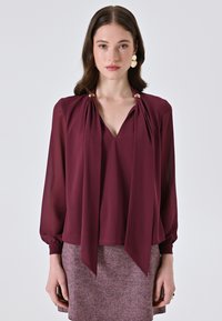 Blusa bordeaux con scollo a V, drappeggi sul davanti legati alle spalle con dettagli dorati e maniche lunghe a sbuffo. Indossata sopra una gonna texturizzata.