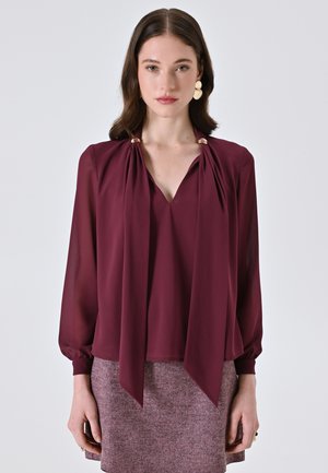 Blusa bordeaux con scollo a V, drappeggi sul davanti legati alle spalle con dettagli dorati e maniche lunghe a sbuffo. Indossata sopra una gonna texturizzata.