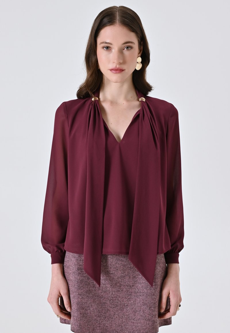 Blusa bordeaux con scollo a V, drappeggi sul davanti legati alle spalle con dettagli dorati e maniche lunghe a sbuffo. Indossata sopra una gonna texturizzata.