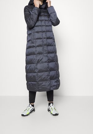 Person, der einen langen schwarzen, gesteppte Daunenmantel mit Kapuze, schwarze Hose und weiße Turnschuhe mit schwarzen und neongelben Akzenten trägt.