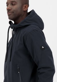 Giacca a vento blu navy con cappuccio, cordini regolabili, tasche con zip e una patch rotonda con logo sulla manica. Tessuto liscio con un design leggero.