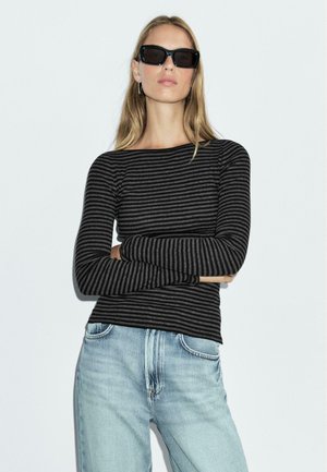 Zwart en grijs gestreepte lange mouwen top met een bateauhals, gecombineerd met lichtblauwe jeans. Model staat met gekruiste armen en draagt zwarte zonnebril.