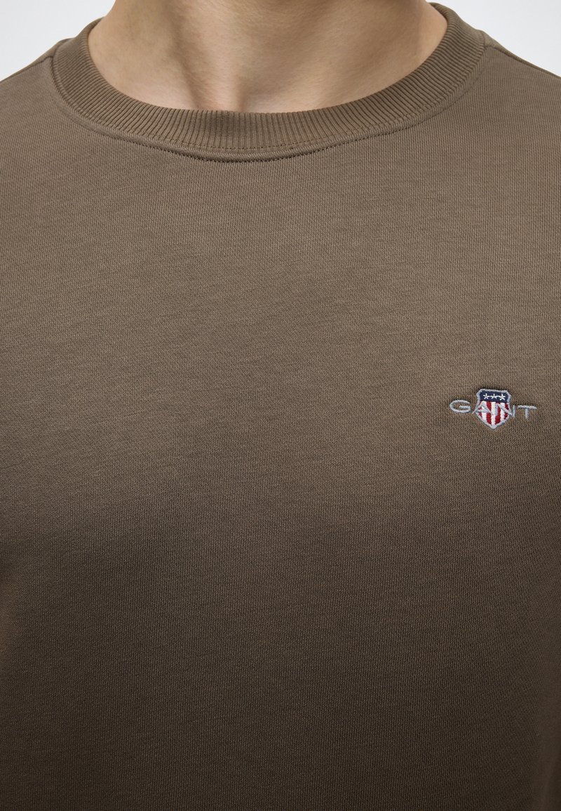 Nærbillede af en person iført en brun crew neck-shirt med et lille GANT-logo og skjoldemblem på venstre bryst.