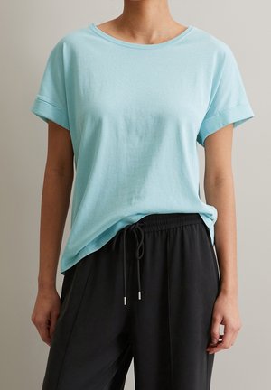 Personne portant un T-shirt bleu clair à manches courtes rentré dans un pantalon noir à cordon, debout devant un fond uni.