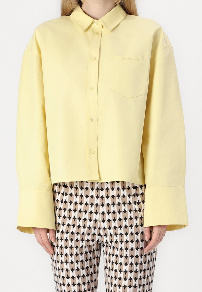 Personne portant une chemise jaune pâle à boutons avec de larges poignets et un pantalon à motifs comportant des formes ovales beige et noires.