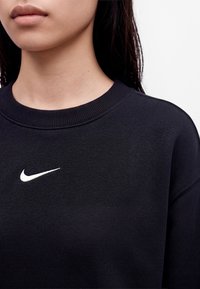 Човек, носещ черен суичър с кръгло деколте и малко бяло лого на Nike Swoosh на гърдите, на едноцветен фон.
