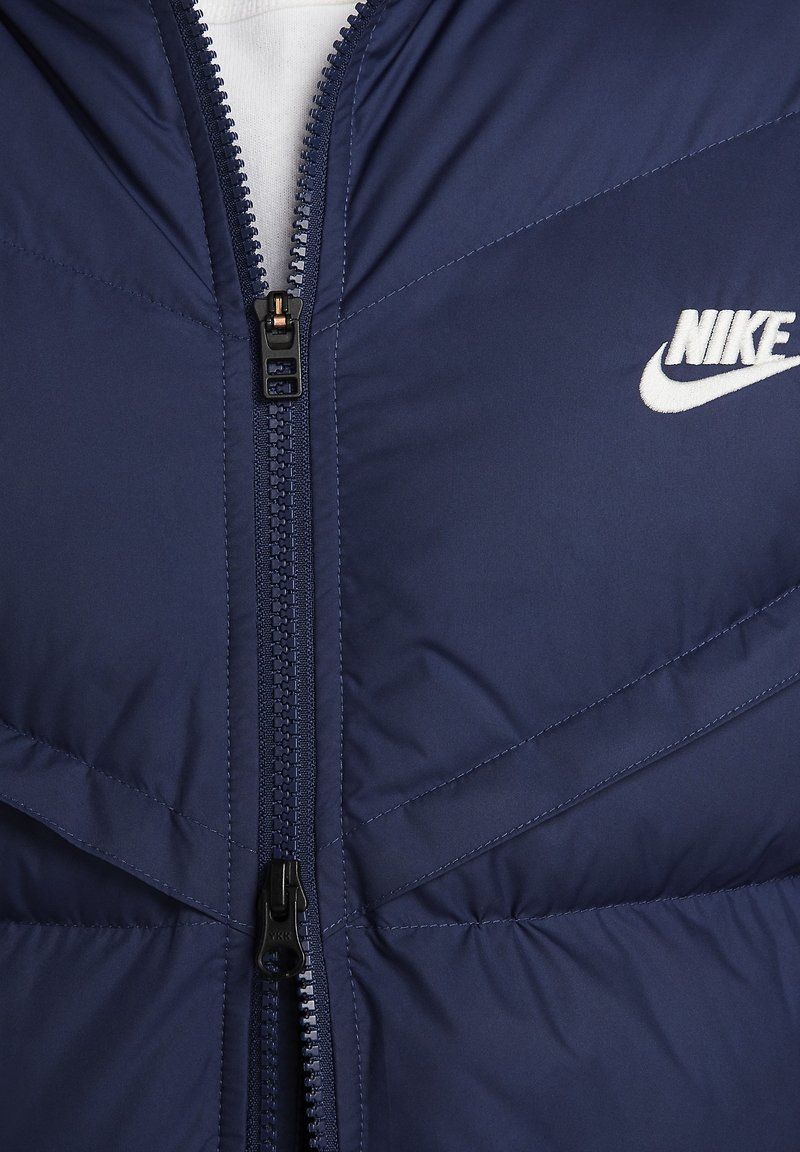 Marineblaue gepolsterte Jacke mit diagonaler Naht, Frontreißverschluss und weißem Nike-Logo. Das Material wirkt glatt und wasserabweisend.