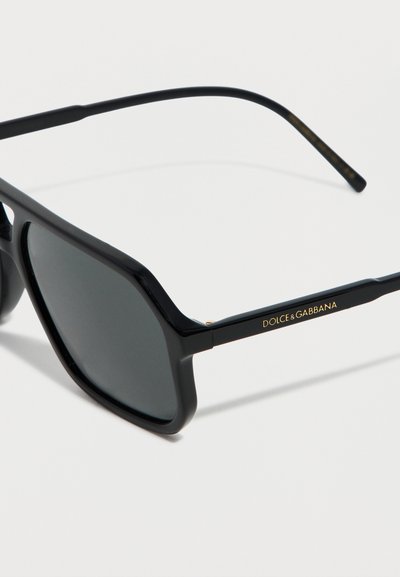 Lunettes de soleil carrées noires avec verres teintés foncés, branches fines noires et logo Dolce & Gabbana doré sur la branche tempes.