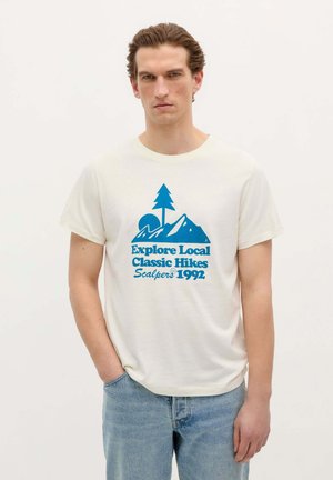 T-shirt in cotone crema con una grafica blu che presenta montagne, un albero e la scritta: "Esplora i classici sentieri locali Scalpers 1992." Vestibilità casual.