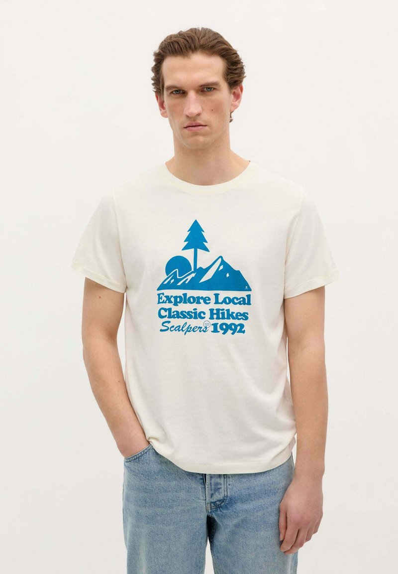 Tricou din bumbac crem, cu un grafic albastru ce prezintă munți, un copac și textul: "Explorează drumețiile clasice locale Scalpers 1992." Croială casual.