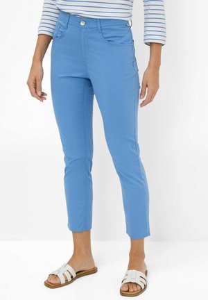 Woman wearing cropped light blue pants, white slip-on sandals, and a white long-sleeve shirt with blue horizontal stripes.