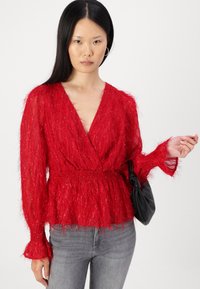Blusa rossa texturizzata con scollatura a V, maniche lunghe e vita arricciata. Presenta una finitura soffice, abbinata a jeans grigi e una borsa nera.