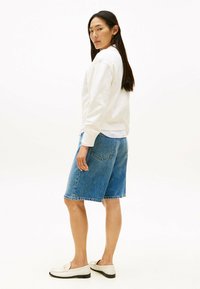 Felpa bianca, pantaloni bermuda di jeans e mocassini bianchi. Dettagli degni di nota includono un colletto a girocollo e tasche posteriori sui pantaloni.