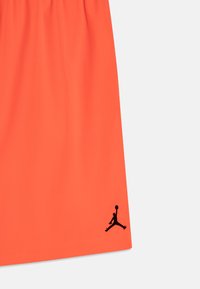 Pantaloni corti arancioni brillanti con cintura elasticizzata e un logo Jordan ricamato nero sull'orlo sinistro inferiore. Tessuto liscio, design minimalista.