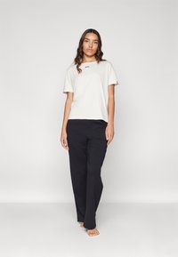 Light beige short-sleeve t-shirt featuring a black logo, paired with loose black trousers. Smooth texture, relaxed fit, and no visible patterns.