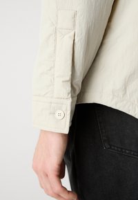 Cream-colored jacket with a textured fabric, featuring a button detail on the cuff and a pocket with a triangular flap.