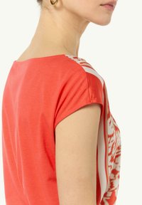 Czerwony top z krótkim rękawem o dekolcie na plecach. Posiada biały wzorzysty akcent z boku. Miękka tkanina o gładkiej fakturze.