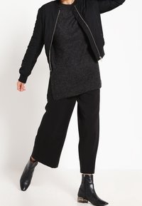 Veste bomber noire, pull oversize gris foncé, pantalon large noir et bottines noires avec un accent métallique. Design simple et moderne.