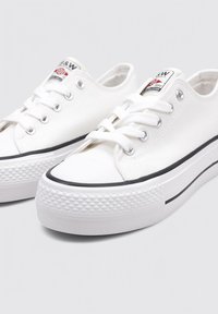 Baskets en toile blanches avec une semelle en caoutchouc, dotées de œillets en métal, de lacets plats blancs et d'une bande noire le long de la semelle. Capuchon de protection texturé.