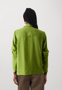Green textured jacket with a collared neckline, long sleeves, and button accents on the cuffs. Worn over brown trousers.