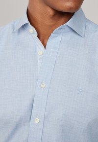 Camicia azzurro chiaro con motivo a scacchi, colletto rigido e dettagli dei bottoni chiari. Presenta un piccolo logo ricamato sul petto.