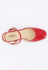 Zapato de charol rojo con punta redondeada, correa en el tobillo y una pequeña hebilla dorada. El interior es beige con una textura suave.