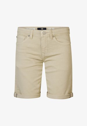 Valkoinen puuvillapaita, jossa on pyöreä logo-ompelus, vaaleanbeige shortsit ja valkoiset korkeavartiset tennarit. Malli poseeraa käsi taskussa.