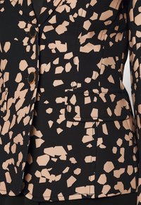 Blazer en tissu noir avec un motif géométrique beige, design ajusté, deux boutons et une poche passepoilée sur le côté gauche.