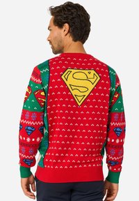 Červený pletený sveter s veľkým žltým logom "S" na chrbte, so zelenými akcentmi a sviatočnými vzormi v modrej a bielej farbe.