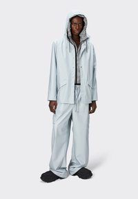 Chaqueta de lluvia impermeable de color azul claro con capucha, botones en la parte delantera y grandes bolsillos, combinada con pantalones cargo de corte holgado a juego. Zapatos texturizados.