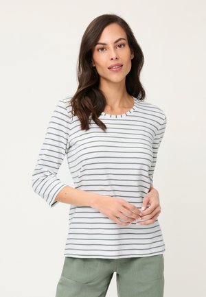 Kobieta o ciemnych włosach, ubrana w białą koszulę z cienkimi szarymi paskami i jasnobeżowe spodnie, stojąca na jednolitym tle.