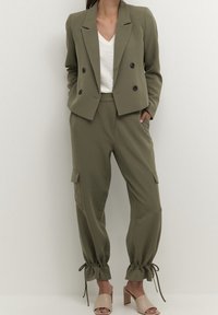 Olivově zelené sako na míru s dvojitým řadovým zapínáním na knoflíky, doplněné olivově zelenými kalhotami ve stylu cargo s nařasenými manžetami a bočními kapsami.