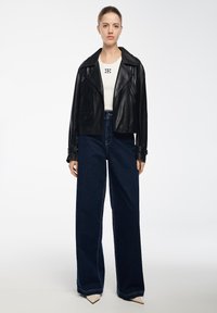 Giacca da biker nera in pelle con zip, t-shirt bianca aderente con logo, e jeans in denim blu a vita alta e gamba larga, completati da tacchi beige a punta.