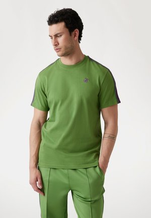 Camiseta de algodón verde con mangas cortas, que presenta rayas moradas a lo largo de los hombros y un pequeño logo en el pecho. Textura suave.