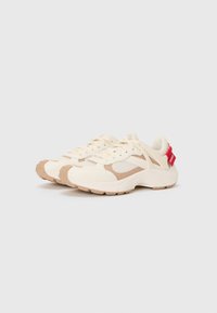 Scarpe sportive beige con tomaia in mesh e materiale sintetico, dotate di suola testurizzata, sistema di chiusura con lacci e un'etichetta rossa sul tallone.